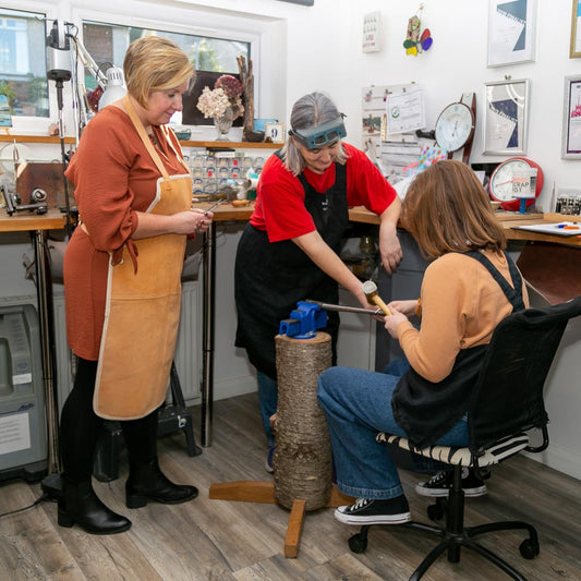 Ruth supervises a seated young guest placing a piece of silver so that it can be hammered into shape. Whilst a lady looks on.