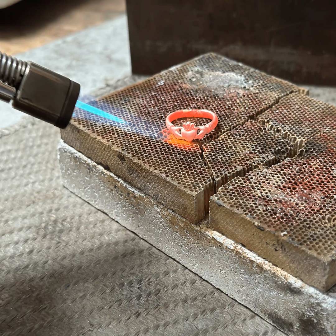 A bright orange-red ring is being held over a torch flame on a metal workbench, with the flame illuminating the Irish Claddagh ring. The scene shows jewelry making in the NI Silver workshop in Holywood, Northern Ireland.