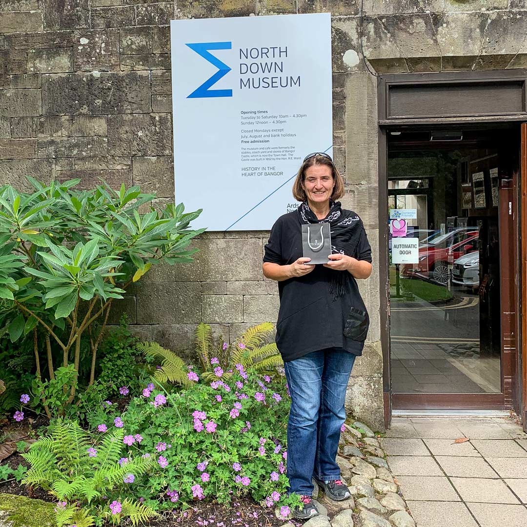 Ruth McEwan-Lyon standing outside the North Down Museum with a NI Silver jewellery gift bag in her hands. Ruth is delivering som emore of our Bangor Bell necklaces which the museum has for sale in their gift shop.