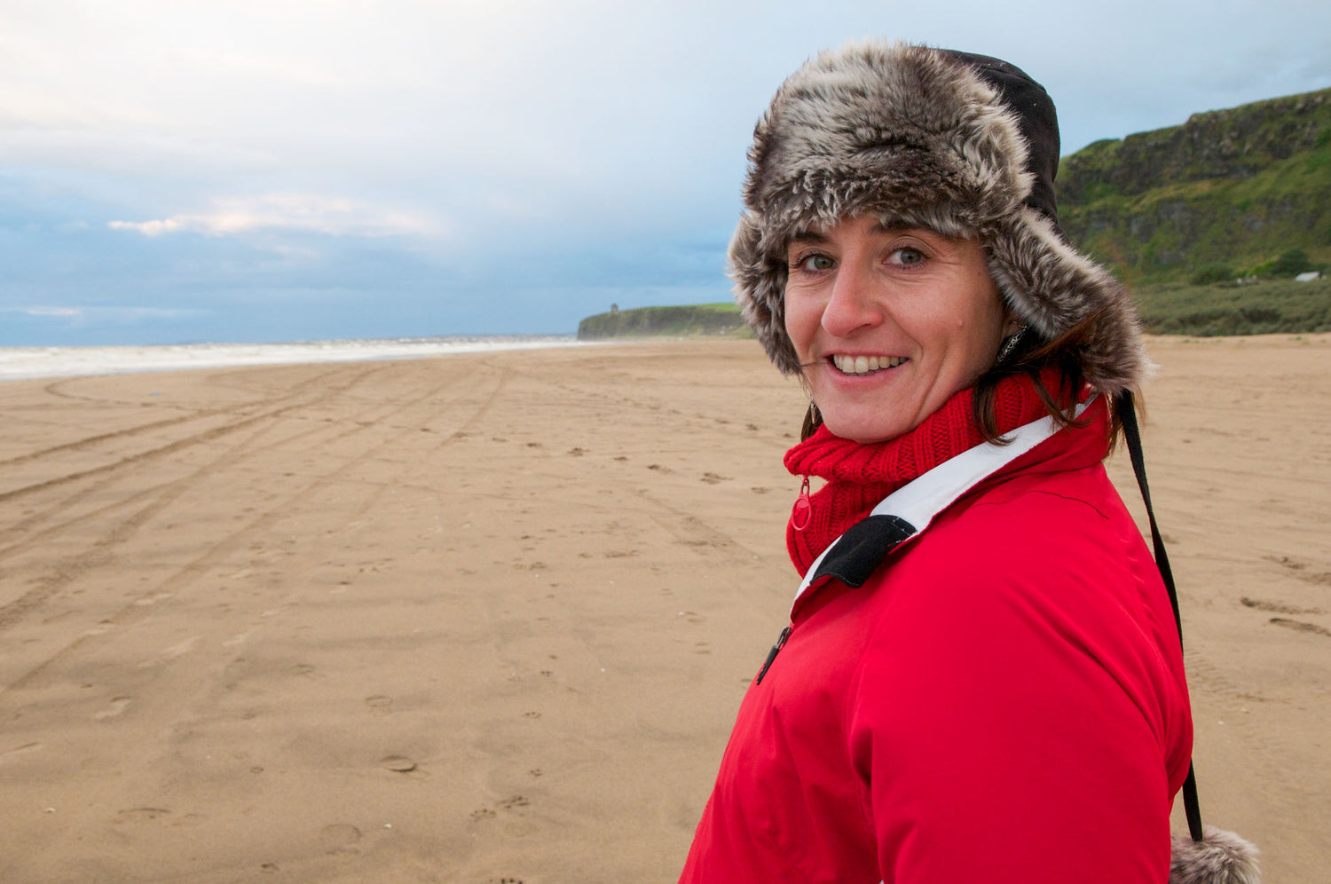 Ruth McEwan-Lyon is NI Silver's Jewellery Maker (Goldsmith).  Ruth is looking straight at the camera and smiling. Ruth is standing on Downhill Strand Beach and wearing a big fluffy hat on a cold day.