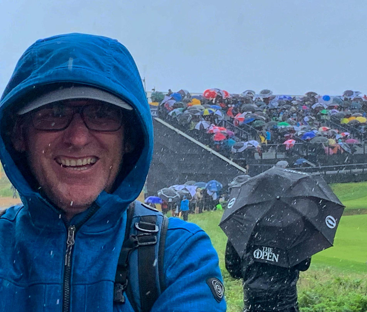 A photo of NI Silver's Steve facing the camera with the British Open golf competition taking part in the background. The rain is pouring down so lots of colourful umbrellas are sheltering people.