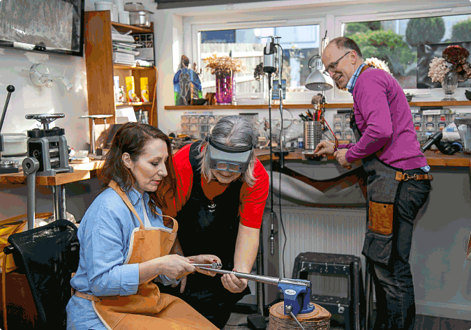 This photo shows the NI Silver Workshop with Ruth leaning over to explain a hammering technique to a seated guest. Steve looks on with a saw in his hand.