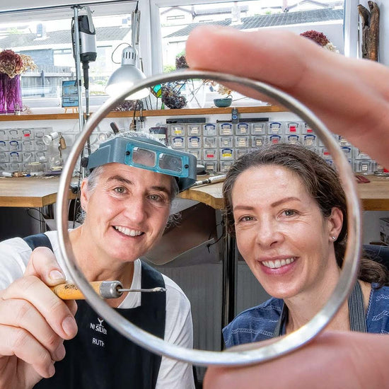 Come to NI Silver Jewellery Making Experience to make your own jewellery.  This image shows two smiling ladies looking at the camera whilst one holds a handmade silver bangle in her hand and the other is pointing at it with a jewellery tool.