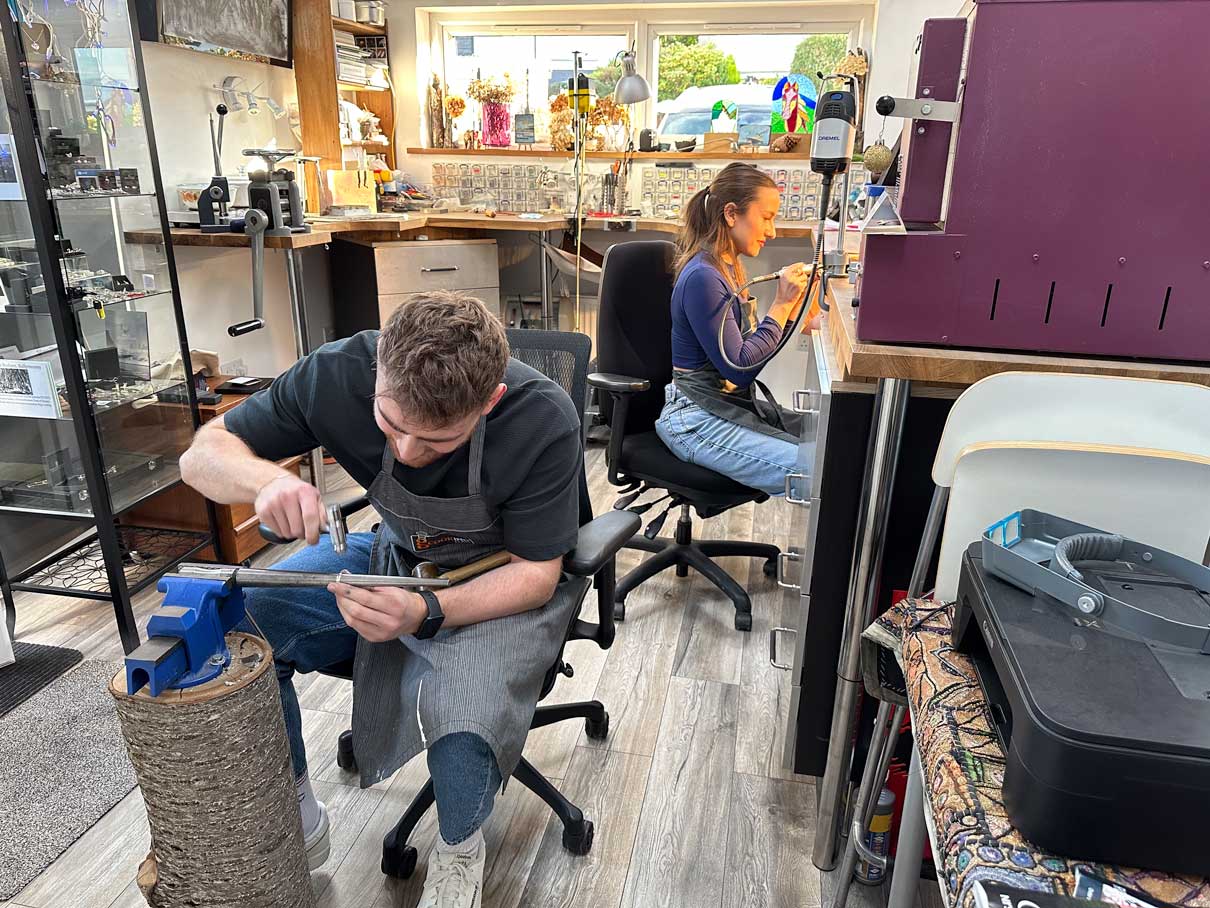 NI Silver's workshop in the Holywood Hills shows a men sat hammering a silver ring on a mandrel and in the background a lady is polishing a ring at a bench.  Both are beginners on a jewellery making workshop with NI Silver.