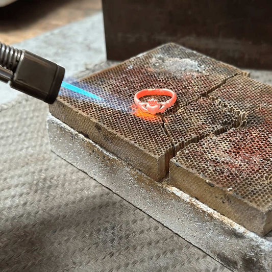 A bright orange-red ring is being held over a torch flame on a metal workbench, with the flame illuminating the Irish Claddagh ring. The scene shows jewelry making in the NI Silver workshop in Holywood, Northern Ireland.  