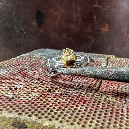 A silver ring with a gold plated crown and heart, resting on top of a rusty metal workbench surface.