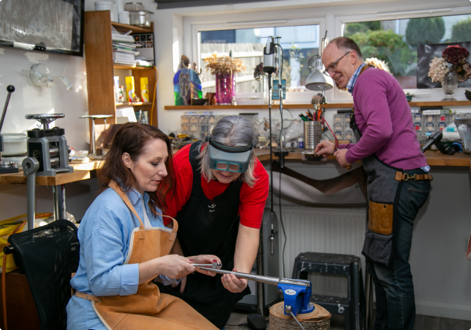 This photo shows the NI Silver Workshop with Ruth leaning over to explain a hammering technique to a seated guest.  Steve looks on with a saw in his hand.
