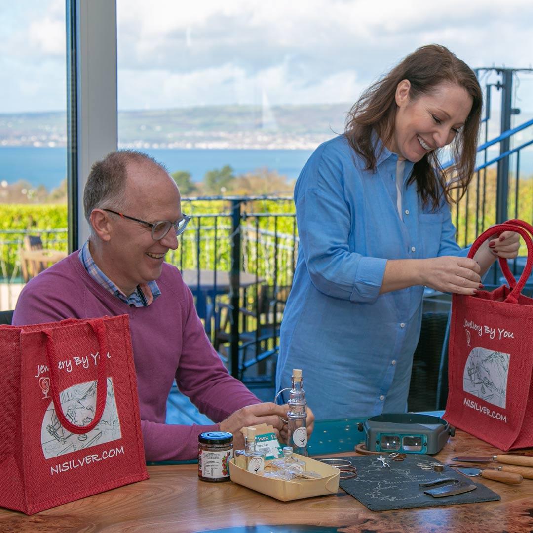 A laughing lady is standing looking into her free NI Silver jute gift bag, whilst a seated laughing man  is looking at a silver bangle. This is the LUX beginners jewellery making experience and you get to take home your jewellery in a gift bag with some Prosecco or Nosecco with some chocolates.